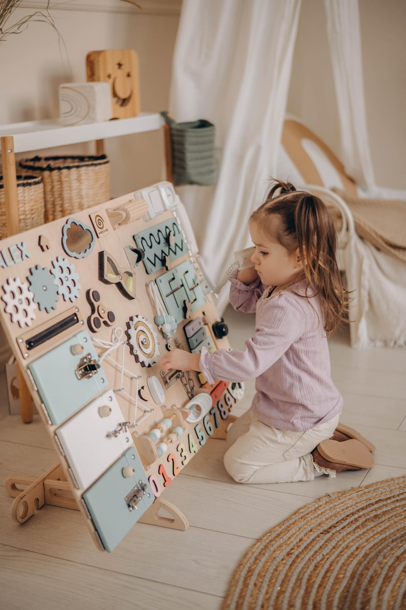 May include: A wooden activity board with various textures and activities for children. The board features a mirror, gears, a zipper, a latch, a maze, a phone, and a number sequence. A child is interacting with the board.