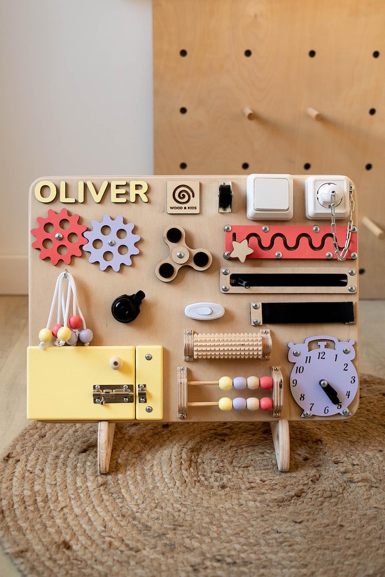 May include: A wooden activity board with various interactive elements for children. Features include gears, a spinner, switches, a clock, and a latch. The board is light brown with colorful accents and the name "OLIVER" in gold letters.