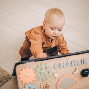 May include: A wooden busy board with colorful gears, a star, a cloud, and the name "CHARLIE" in blue letters. A baby in an orange outfit is interacting with the board. The board is designed for sensory and fine motor skill development.