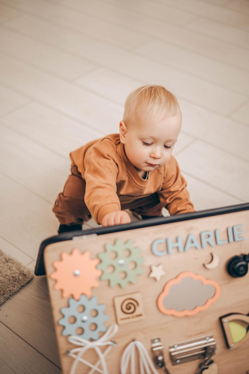 May include: A wooden busy board with colorful gears, a star, a cloud, and the name "CHARLIE" in blue letters. A baby in an orange outfit is interacting with the board. The board is designed for sensory and fine motor skill development.