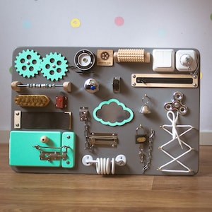 May include: A gray wooden busy board with various interactive elements for children. Features include gears, switches, a zipper, a bell, a spinner, a brush, a latch, and a shoelace tying activity. The board is approximately 24 inches wide and 18 inches tall.