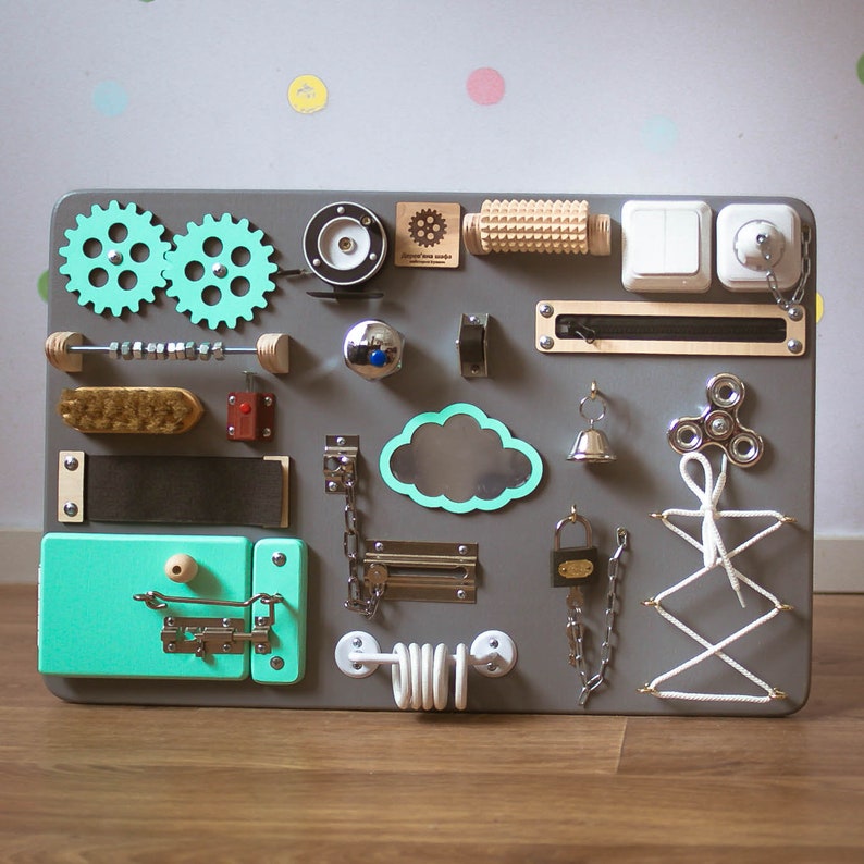 May include: A gray wooden busy board with various interactive elements for children. Features include gears, switches, a zipper, a bell, a spinner, a brush, a latch, and a shoelace tying activity. The board is approximately 24 inches wide and 18 inches tall.