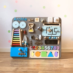 May include: A gray wooden activity board with various interactive elements for children. Features include gears, a ladder, an abacus, a maze, and shape sorters. The name "Archie" is displayed in blue letters. Includes a variety of colors and textures.