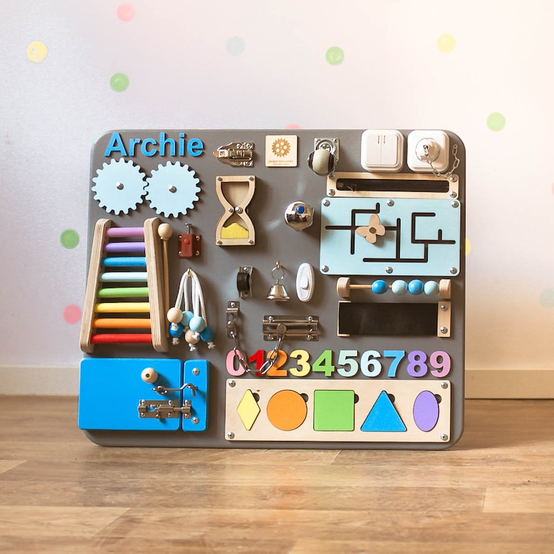 May include: A gray wooden activity board with various interactive elements for children. Features include gears, a ladder, an abacus, a maze, and shape sorters. The name "Archie" is displayed in blue letters. Includes a variety of colors and textures.