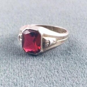 May include: A silver-toned ring featuring a large, rectangular, faceted deep red gemstone set in the center. The band has a smooth, polished finish with structural metal detailing on the sides flanking the central stone setting.