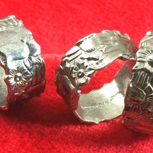 May include: Three silver-toned napkin rings with a scalloped edge and a raised floral pattern embossed on the exterior surface. The metal bands feature a continuous design of small flowers and leaves, with a reflective finish and a hollow center.