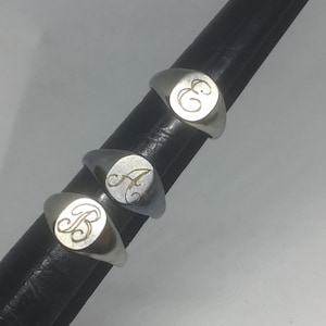 May include: Three silver-toned signet rings arranged vertically on a black ring mandrel. Each ring features a smooth, polished surface with a single engraved letter in a cursive script. The letters visible on the rings are B, A, and E.