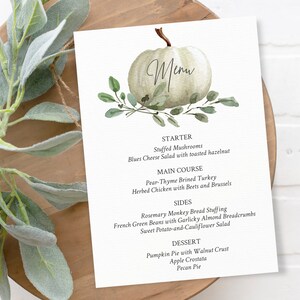 May include: A white pumpkin with green leaves and the word "Menu" written on it. The menu lists a starter, main course, sides, and dessert. The dishes include Stuffed Mushrooms, Pear-Thyme Brined Turkey, Rosemary Monkey Bread Stuffing, and Pumpkin Pie with Walnut Crust.