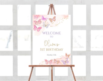 May include: A welcome sign for Olive's 1st birthday, displayed on a wooden easel. The sign features watercolor butterflies in pink, purple, and gold, with the text "Welcome to Olive's 1st Birthday" in gold and purple. The date is October 15th.