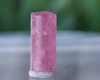 May include: A pink and white crystal specimen with a striated texture. The crystal is sitting on a white surface.