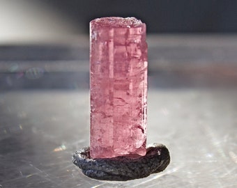 May include: A pink, translucent, cylindrical crystal with striations on its surface. The crystal is mounted on a dark gray base.
