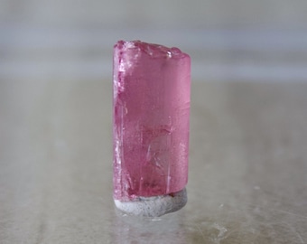 May include: A pink, translucent crystal with a rough, natural texture. The crystal is shaped like a prism and is sitting on a white, textured surface.