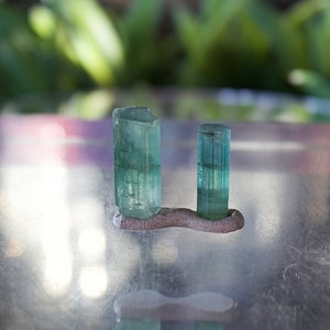 May include: Two green gemstones, likely emeralds, are displayed on a small, gray, clay stand. The gemstones are cut into rectangular prisms and have a translucent, glassy appearance.
