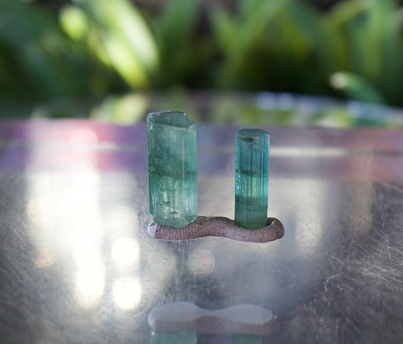 May include: Two green gemstones, likely emeralds, are displayed on a small, gray, clay stand. The gemstones are cut into rectangular prisms and have a translucent, glassy appearance.