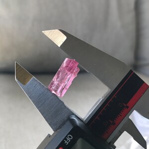 May include: A pink tourmaline crystal is measured with a caliper. The crystal is approximately 1.5 inches long.
