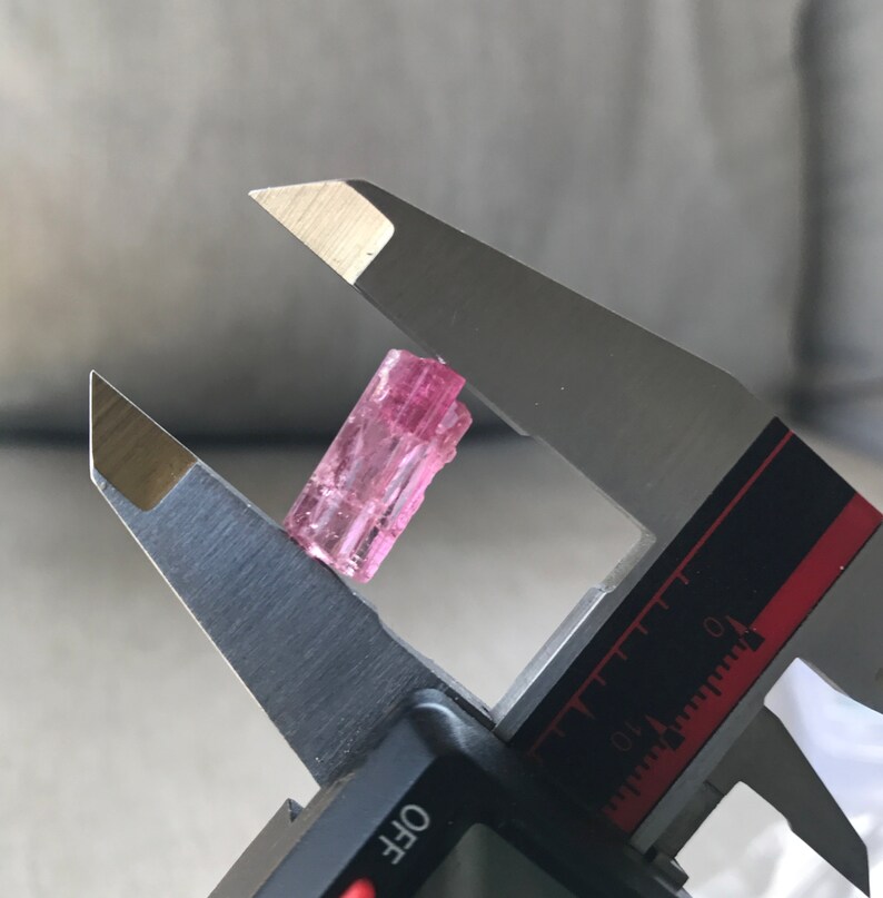 May include: A pink tourmaline crystal is measured with a caliper. The crystal is approximately 1.5 inches long.