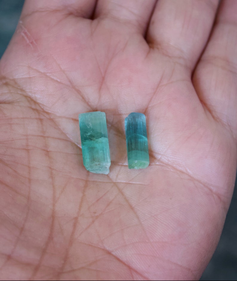 May include: Two green aquamarine crystals, one slightly larger than the other, are shown in a hand. The crystals are translucent and have a light green color.