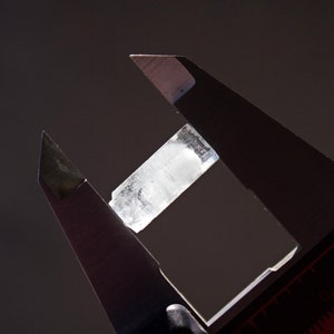 Pode incluir: Um cristal transparente est&aacute; sendo medido com um paqu&iacute;metro de prata. O cristal tem cerca de 5 cm de comprimento.