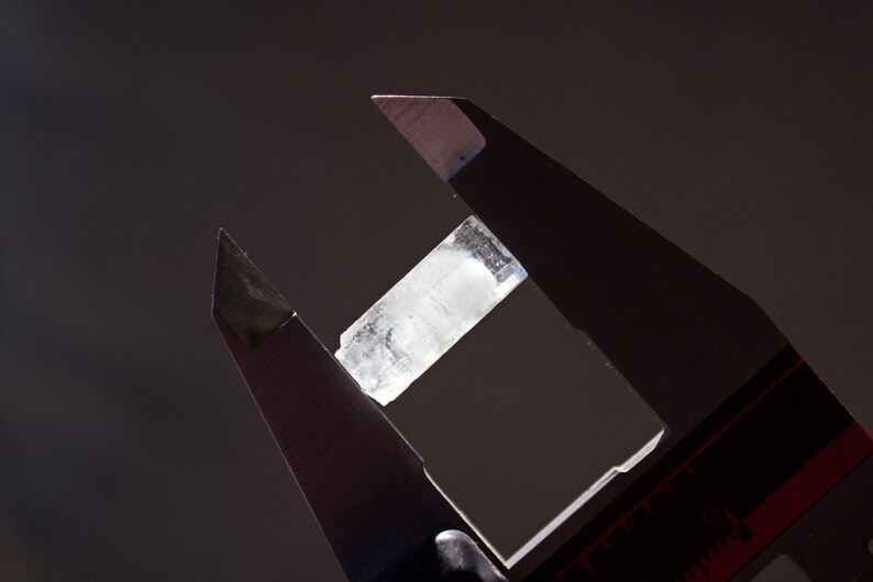 Pode incluir: Um cristal transparente est&aacute; sendo medido com um paqu&iacute;metro de prata. O cristal tem cerca de 5 cm de comprimento.