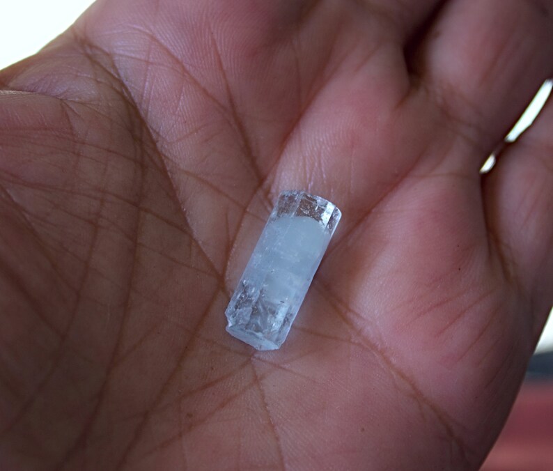 Pode incluir: Um cristal de &aacute;gua-marinha transparente e azul claro com forma retangular. O cristal tem aproximadamente 5 cm de comprimento e est&aacute; na palma da m&atilde;o.