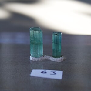 May include: Two green gemstone crystals on a gray display stand. The crystals are long and slender, with a slightly rounded top. The stand is made of a rough, textured material.