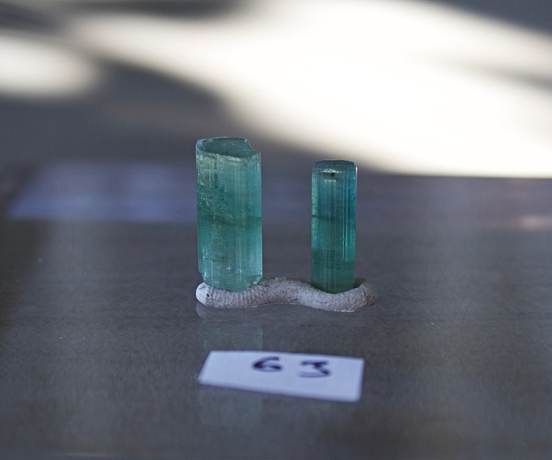 May include: Two green gemstone crystals on a gray display stand. The crystals are long and slender, with a slightly rounded top. The stand is made of a rough, textured material.