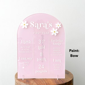 K&ouml;nnte beinhalten: Ein rosa Acryl-Meilenstein-Schild f&uuml;r das erste Jahr mit wei&szlig;em Text und G&auml;nsebl&uuml;mchen-Akzenten. Das Schild enth&auml;lt den Namen "Sara's", eine Gr&ouml;&szlig;e von 84 cm, 24 Pfund und Lieblingsspeisen. Es wird auf einem Holzst&auml;nder ausgestellt.