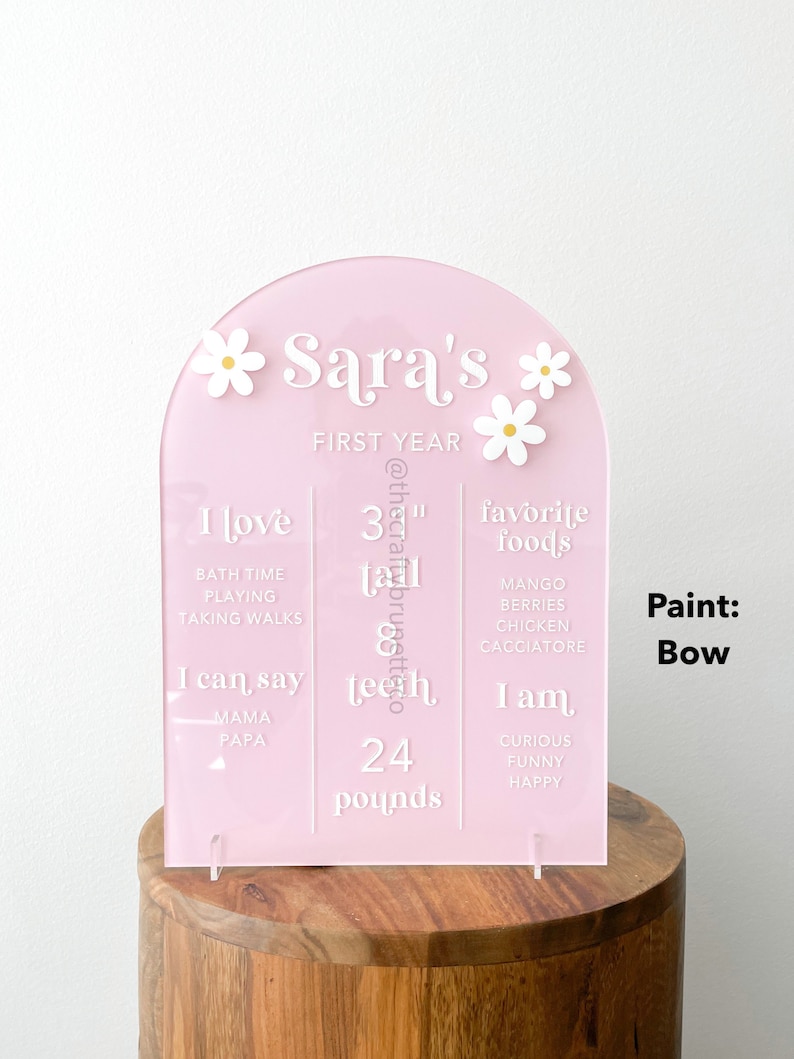 K&ouml;nnte beinhalten: Ein rosa Acryl-Meilenstein-Schild f&uuml;r das erste Jahr mit wei&szlig;em Text und G&auml;nsebl&uuml;mchen-Akzenten. Das Schild enth&auml;lt den Namen "Sara's", eine Gr&ouml;&szlig;e von 84 cm, 24 Pfund und Lieblingsspeisen. Es wird auf einem Holzst&auml;nder ausgestellt.