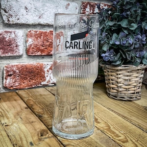 May include: A clear glass beer pint glass with the word "CARLING" in white and black lettering. The glass has a ribbed design and is set against a brick wall and wooden surface.