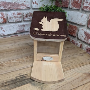 Puede incluir: Comedero de madera para ardillas con un techo marrón y un frente de plástico transparente. El techo tiene una silueta de una ardilla con el texto "All squirrels welcome".