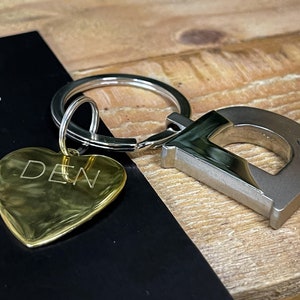 May include: A gold heart-shaped keyring with the word "DEN" engraved on it. The keyring is attached to a silver key ring with a silver letter "D" charm with two small rhinestones.