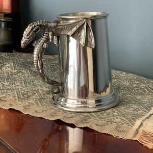 May include: A silver metal beer stein with a dragon handle. The dragon is detailed with scales and wings.