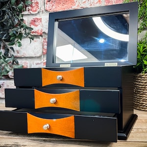 May include: A black wooden jewellery box with three drawers and a hinged lid with a mirror. The drawers have orange wood fronts with silver knobs.