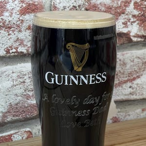 May include: A pint glass filled with dark stout, topped with a creamy, tan head. The Guinness logo and a harp are visible on the glass. Text on the glass reads "A lovely day for Guinness Dad Love Beth."