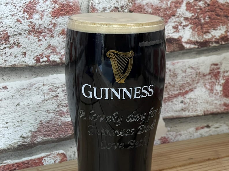 May include: A pint glass filled with dark stout, topped with a creamy, tan head. The Guinness logo and a harp are visible on the glass. Text on the glass reads "A lovely day for Guinness Dad Love Beth."