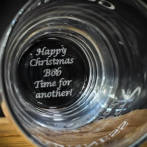 May include: Clear glass with the text "Happy Christmas Bob Time for another!" etched on the bottom. The glass has a wide rim and a cylindrical shape. The image is taken from a low angle, looking up into the glass.