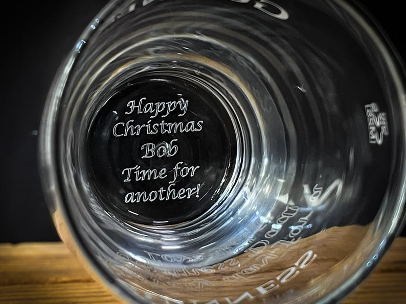 May include: Clear glass with the text "Happy Christmas Bob Time for another!" etched on the bottom. The glass has a wide rim and a cylindrical shape. The image is taken from a low angle, looking up into the glass.