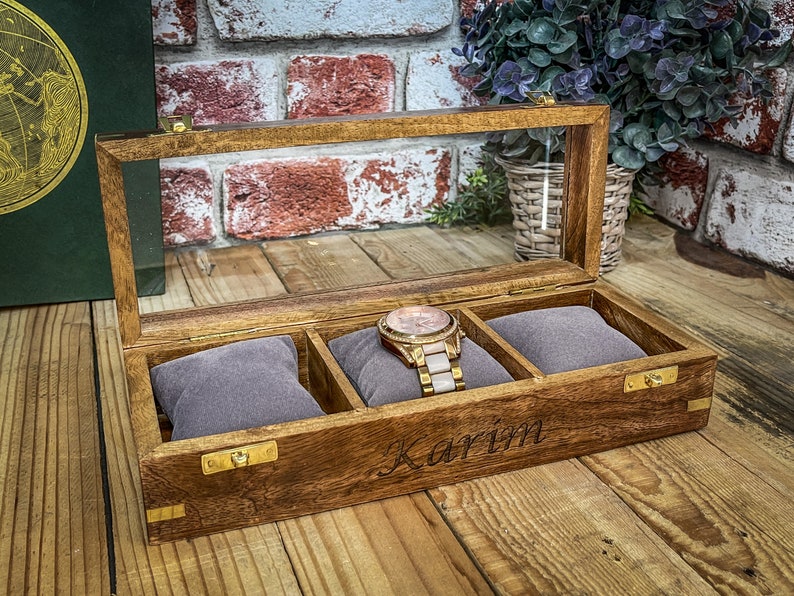 May include: A wooden watch box with a glass top and three compartments. The box is engraved with the name "Karim".