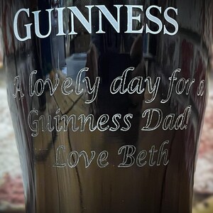 May include: A black Guinness pint glass with white lettering. The text includes "GUINNESS", "A lovely day for a Guinness Dad", and "Love Beth". The glass has a curved shape and is likely made of glass.