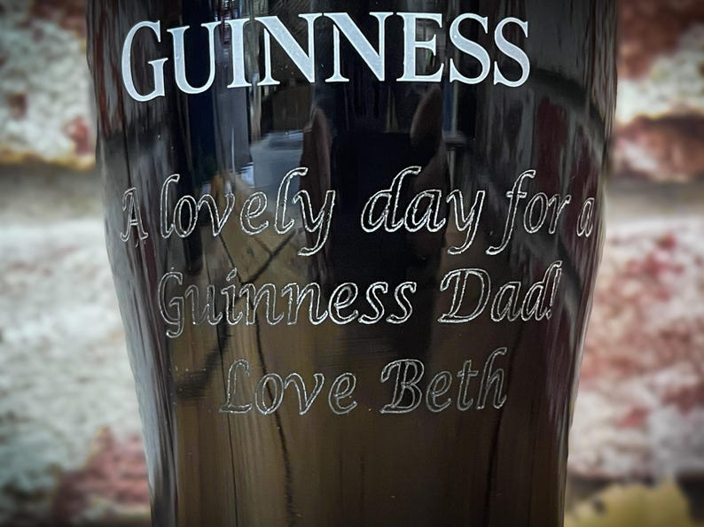 May include: A black Guinness pint glass with white lettering. The text includes "GUINNESS", "A lovely day for a Guinness Dad", and "Love Beth". The glass has a curved shape and is likely made of glass.