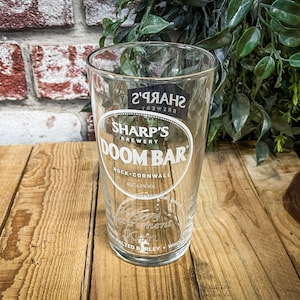 May include: A clear glass pint glass with a white logo that says "Sharp's Brewery Doom Bar Rock - Cornwall Alc 4.0% Vol.  Malt Barley, Wheat, Hops"
