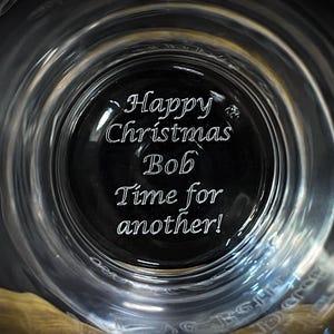 May include: A clear glass with the words "Happy Christmas Bob Time for another!" etched on the bottom. The glass has a wide rim and a dark center, creating a unique visual effect. The text is in a cursive font.
