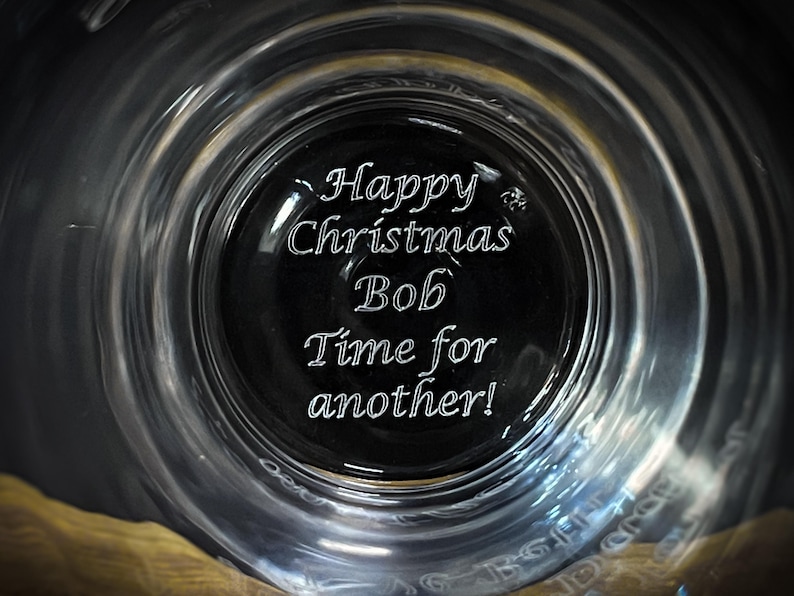 May include: A clear glass with the words "Happy Christmas Bob Time for another!" etched on the bottom. The glass has a wide rim and a dark center, creating a unique visual effect. The text is in a cursive font.