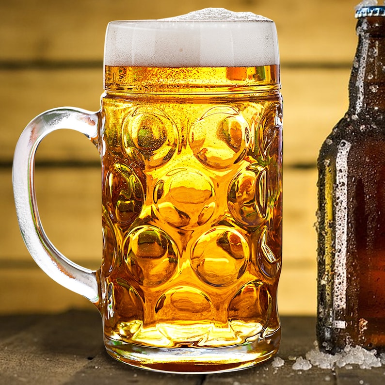 May include: A large, clear glass beer stein filled with golden beer and a thick white head of foam. A brown beer bottle is next to the stein. The stein has a handle and a textured pattern. The background is a wooden surface.