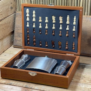 Könnte beinhalten: Eine Holzkiste mit einem Scharnierdeckel, die ein Schachspiel, zwei Schnapsgläser und eine Edelstahl-Schnapsflasche enthält. Die Flasche hat die Gravur "VIP".