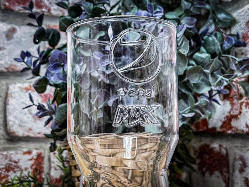 May include: A clear glass with the Pepsi Max logo etched on the side. The glass is sitting on a woven surface.