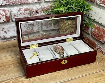May include: A cherry-stained wooden watch box with a glass top, featuring four compartments with white cushions. A gold-toned watch and key are visible inside. The box has gold-coloured hinges and a lock, and is displayed on a wooden surface.