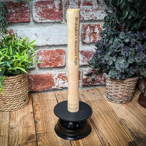 Op de afbeelding: Een lichtbruine houten stamhouder met de tekst "Brandon's massive logs" gegraveerd op de zijkant. De houder staat op een zwarte basis. De achtergrond bevat planten in gevlochten manden en een bakstenen muur.