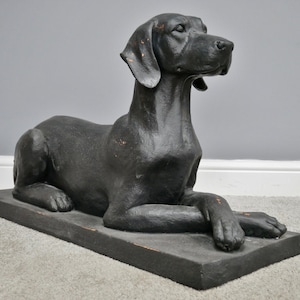 May include: A black dog statue in a reclining pose, resting on a rectangular base. The dog has a sleek, dark coat with visible details, including its ears, snout, and paws. The statue is made of a solid material, and the overall design is classic and elegant.