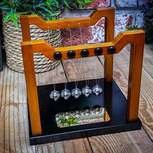 Op de afbeelding: Een houten Newtons wieg met vijf zilveren ballen die aan touwtjes hangen. De ballen zijn opgehangen aan een houten frame met een zwarte basis. De basis heeft een spiegelend oppervlak dat de ballen reflecteert.